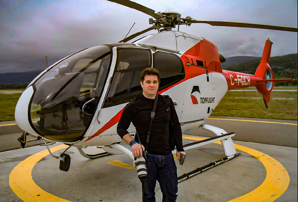 Shaun Kennedy, Penticton photographer and creative marketer, standing with his camera beside a helicopter on a helipad in British Columbia