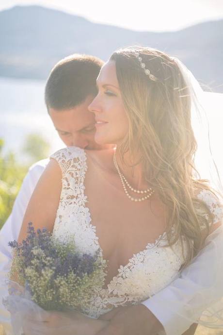 Sunlit close-up of bride in lace dress with groom embracing her from behind beside the lake