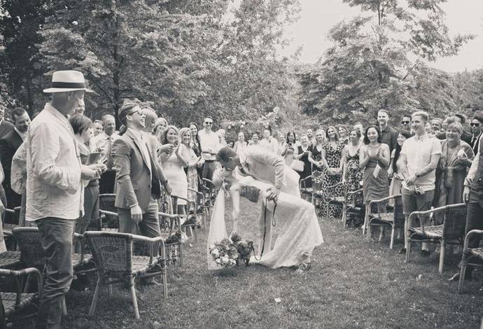 Black-and-white wedding ceremony photograph of newlyweds greeting their dog as guests smile and applaud