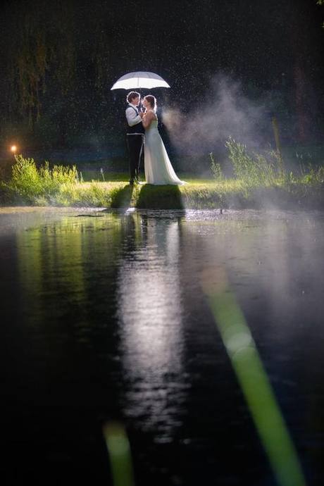Newlyweds holding a glowing umbrella in the rain beside water at night