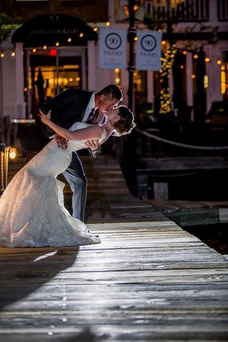 Bride and groom dipping into a kiss on a wooden dock beside twinkling lights at night