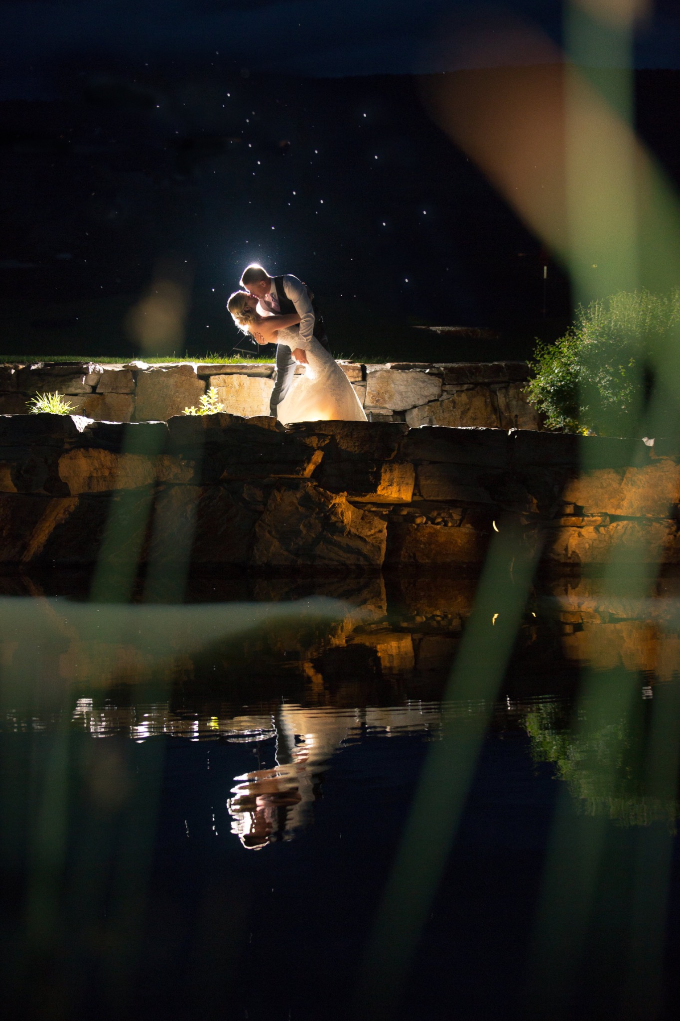 Newlyweds sharing a dramatic nighttime embrace beside a stone terrace with their reflection in the water at a Kelowna wedding