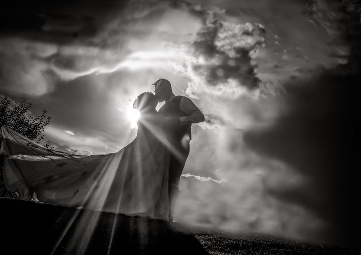 Black-and-white silhouette of a newlywed couple kissing against dramatic clouds and sunbeams with the bride’s wedding dress flowing behind her