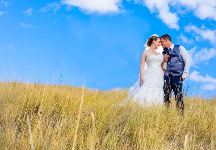 Bride and groom standing close together among tall golden grasses beneath an expansive blue sky