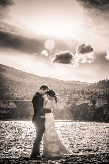 Warm sepia-toned wedding portrait of a couple embracing on a lakeshore with mountains behind them