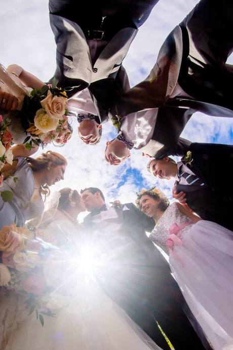 Wedding party gathered in a circle around the newlyweds, photographed from below against the bright sky