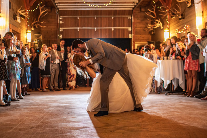 Newlyweds share a dramatic dip kiss on the dance floor as their wedding guests watch inside a warmly lit rustic reception venue