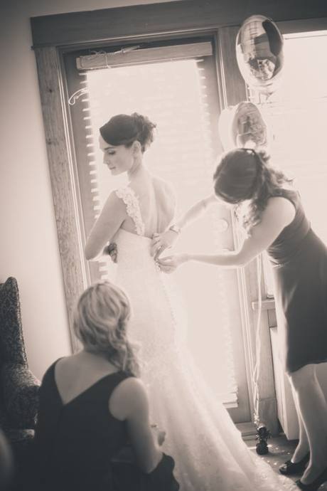Black-and-white documentary wedding photograph of a bride having her dress fastened
