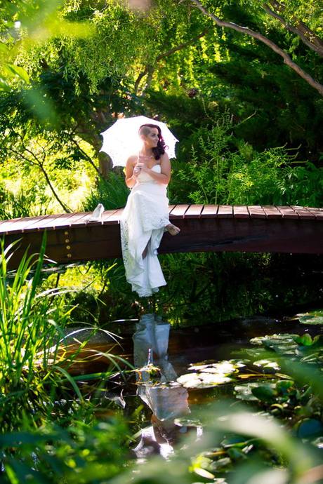 Bride holding a white parasol while sitting on a wooden bridge over a garden pond