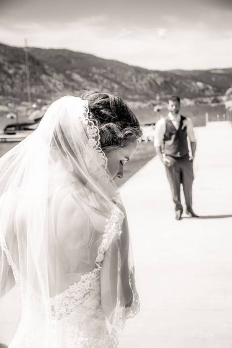 Black-and-white portrait of a veiled bride by the water with a groom farther along the dock